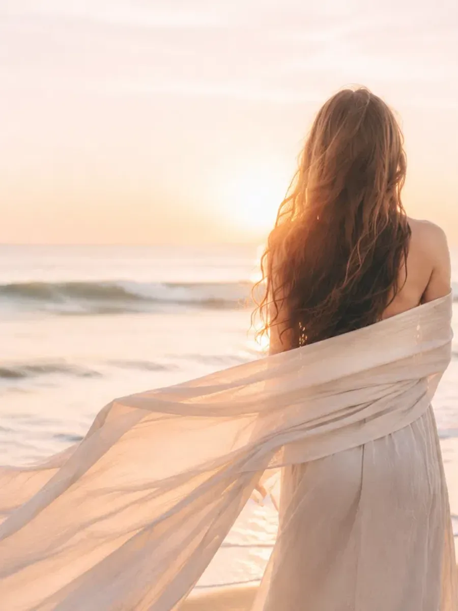 A woman in a flowing dress standing at the shoreline at sunrise